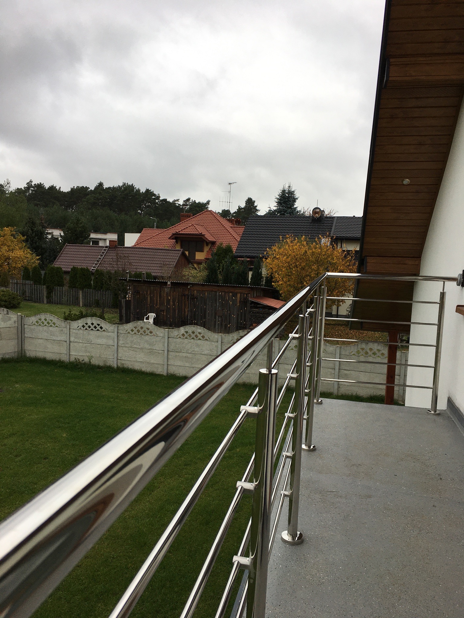 Balkon z polerowaną, metalową balustradą o minimalistycznym designie, z widokiem na trawnik, ogrodzenie z prefabrykatów betonowych i zabudowę jednorodzinną w tle, pochmurne niebo.