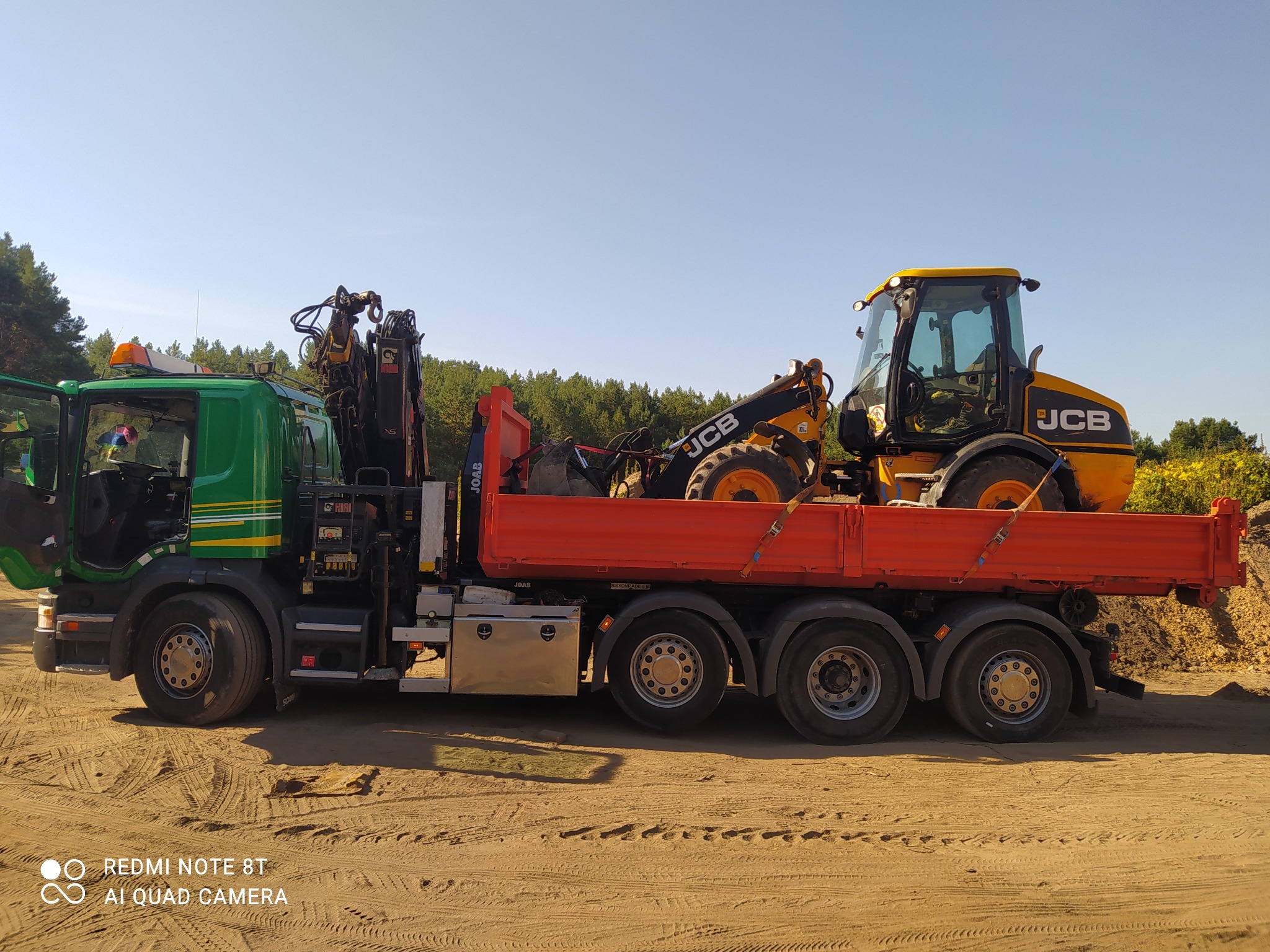 Zielony samochód ciężarowy z dźwigiem przewożący żółtą koparko-ładowarkę JCB na pomarańczowej platformie, widok z boku na piaszczystym terenie.