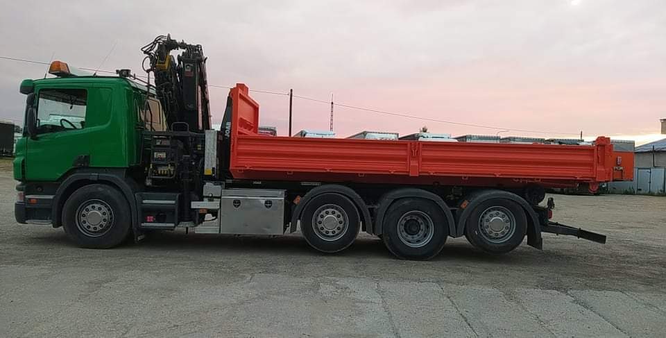 Zielono-pomarańczowa ciężarówka z żurawiem i wywrotką JOAB na placu, widok z boku, zachodzące słońce w tle.