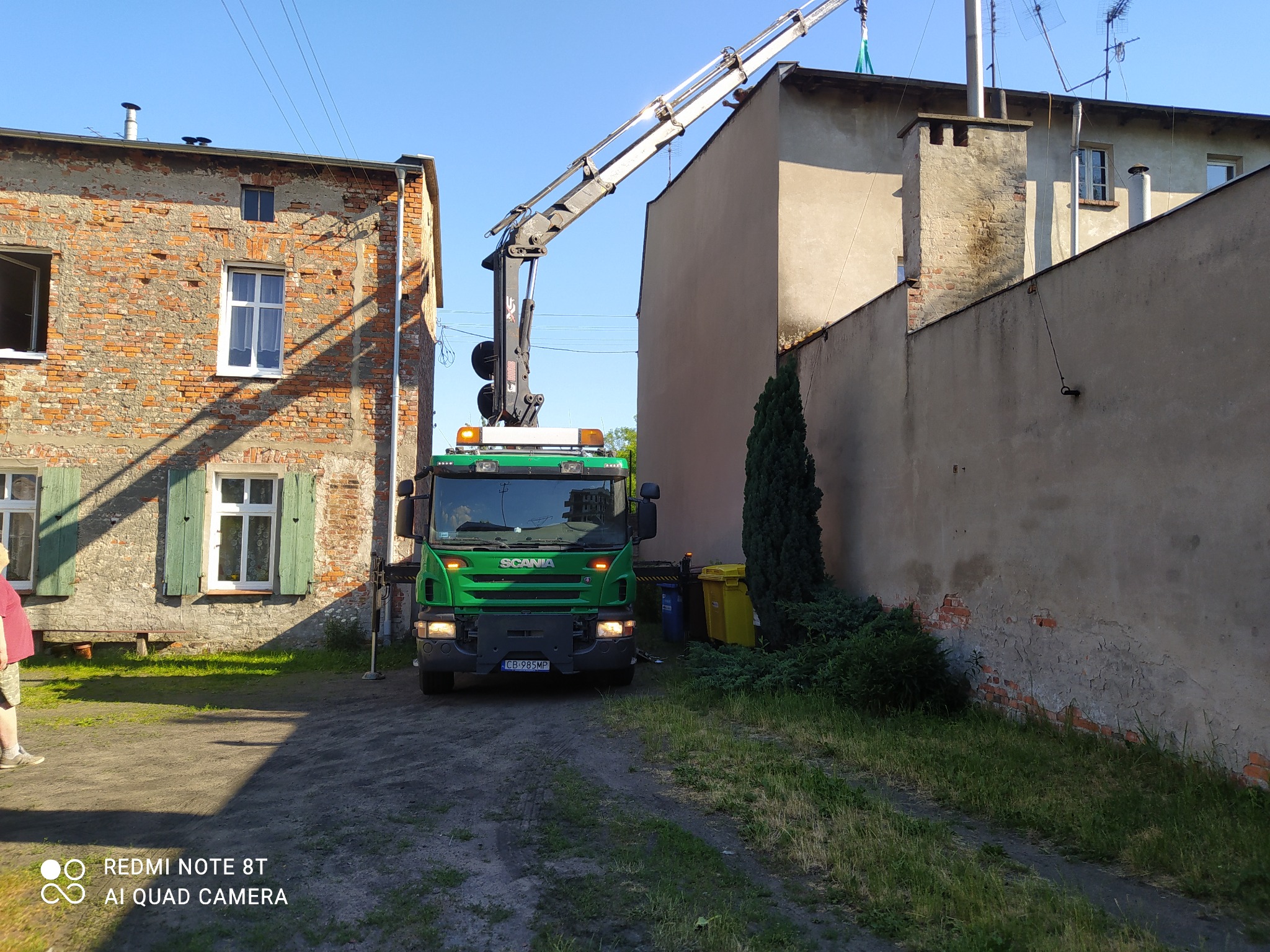 Zielony samochód ciężarowy Scania z dźwigiem ustawiony pomiędzy budynkami, prawdopodobnie podczas prac na wysokościach.