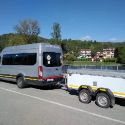Srebrny bus z przyczepką na tle górskiego krajobrazu. Bus posiada ciemne szyby i żółty pas. Przyczepka biała z szarą plandeką. Błękitne niebo w tle.