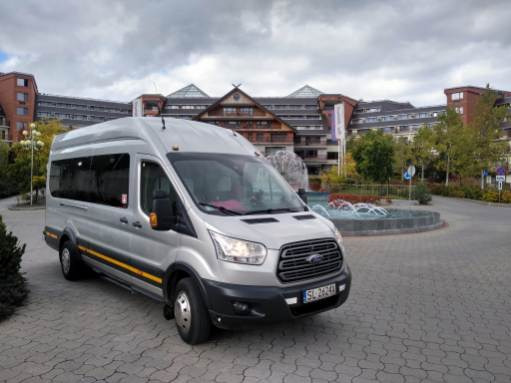 Srebrny bus Ford Transit z żółtymi pasami parkuje na brukowanym placu przed hotelem z fontanną w tle.