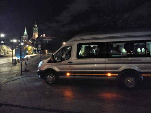 Srebrny bus zaparkowany nocą na tle Wawelu, widoczne światła miejskie i zarys zamku.