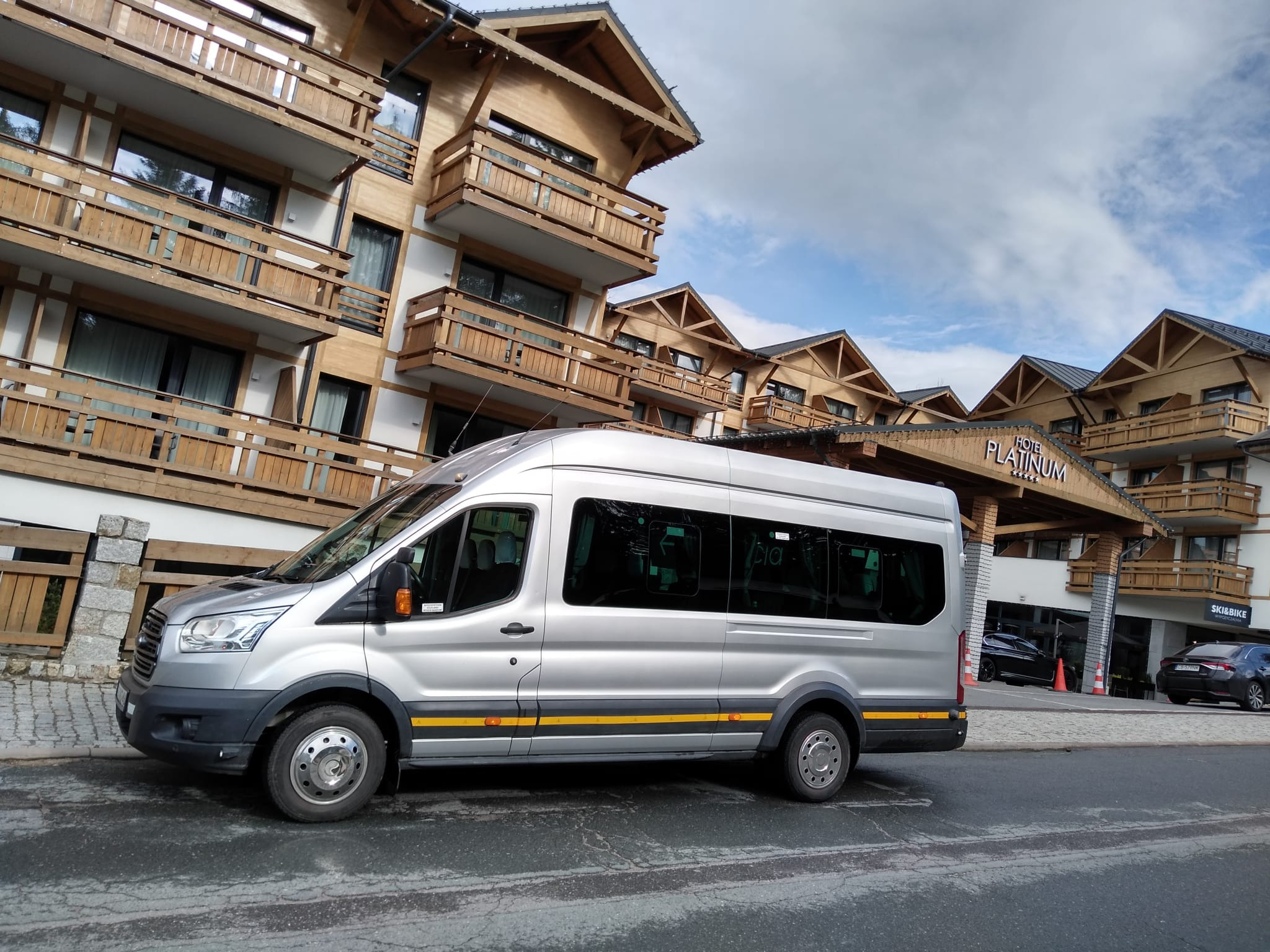 Srebrny bus Ford Transit z żółtymi akcentami na tle drewnianego hotelu Platinum z balkonami i podjazdem.