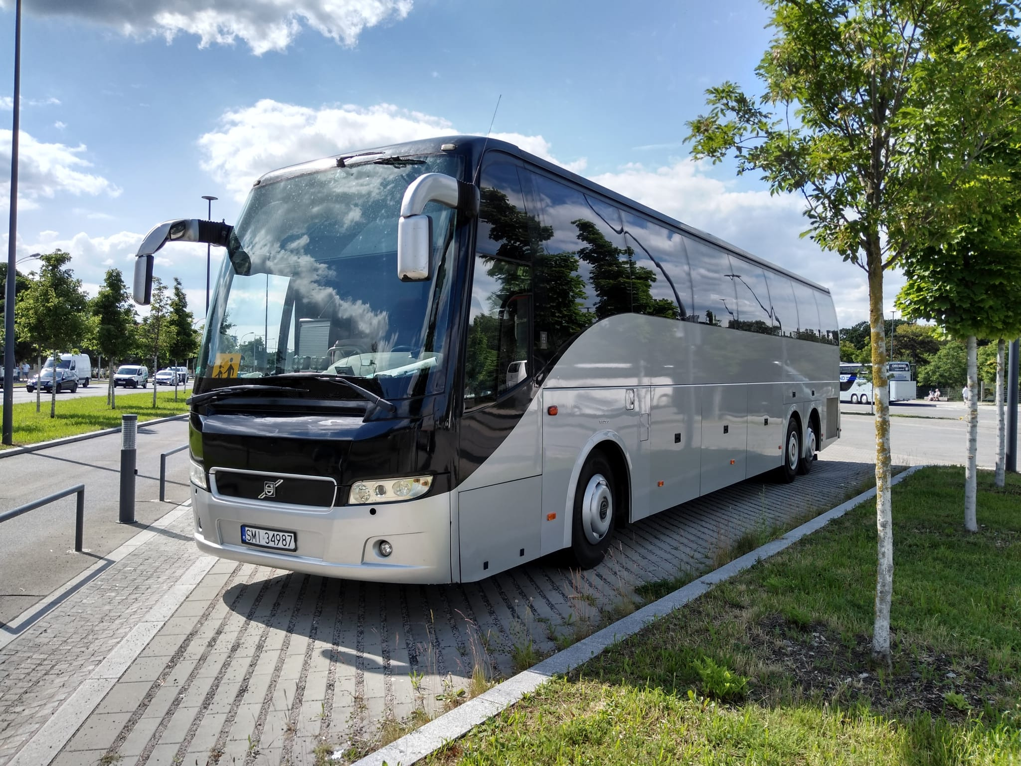Srebrny autokar Volvo z czarnym przodem, zaparkowany na kostce brukowej obok trawnika z młodymi drzewkami, numer rejestracyjny SMI 34987.