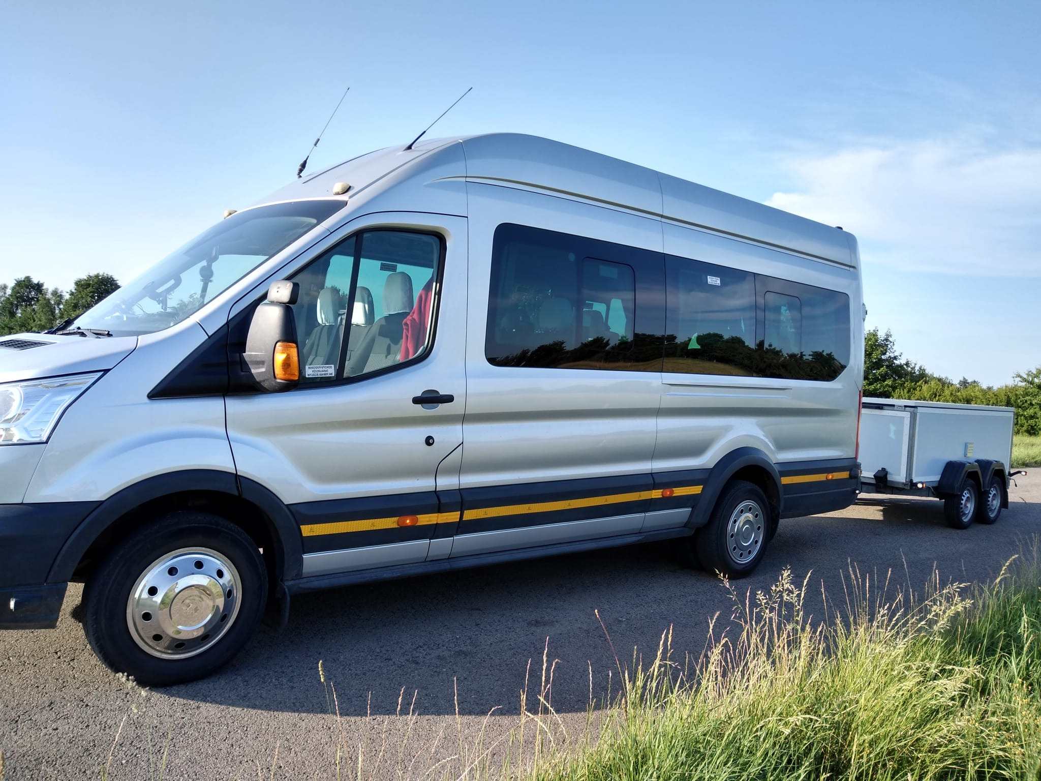 Srebrny bus z przyczepą na asfalcie, widok z boku, antena na dachu, żółte pasy odblaskowe na dole pojazdu.