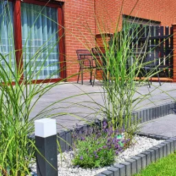 Nowoczesny taras z szarej kostki brukowej, otoczony ozdobnymi trawami i lawendą w białym żwirku, z widokiem na ceglany dom i czarne meble ogrodowe.