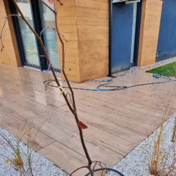 Nowoczesny taras z płytek imitujących drewno, otoczony białym żwirem i zielenią, z widocznym fragmentem budynku w tle.