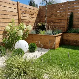 Nowoczesny ogród z drewnianym ogrodzeniem, podwyższonymi rabatami, białym oświetleniem kulistym i ozdobnymi trawami na tle błękitnego nieba.