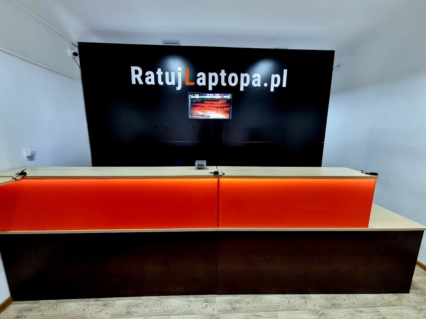 Recepcja serwisu komputerowego z podświetlanym na pomarańczowo blatem i logo firmy na czarnym tle.