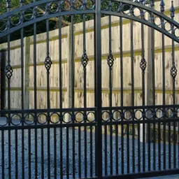 Czarna, metalowa brama z dekoracyjnymi elementami na tle drewnianego ogrodzenia.