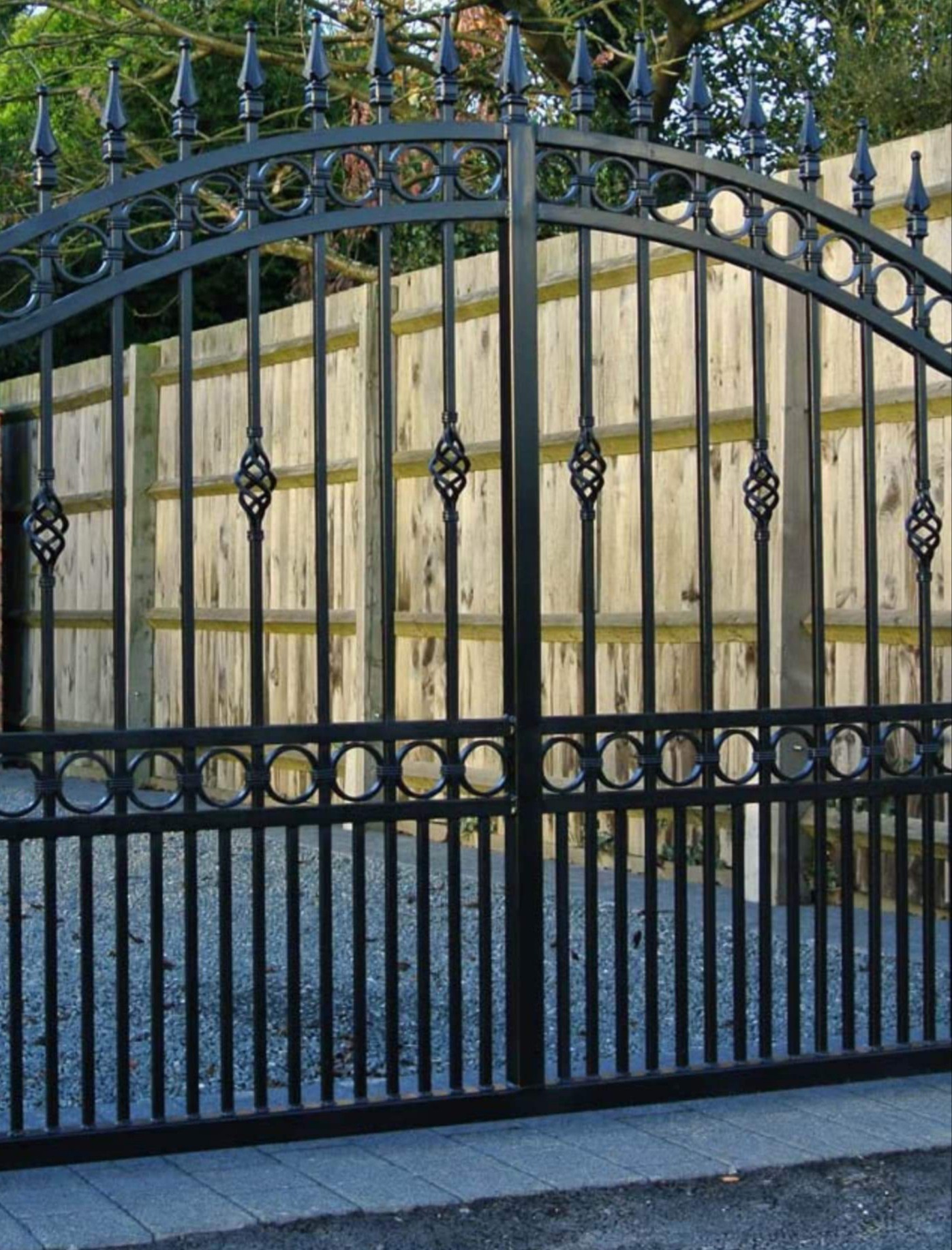 Czarna, metalowa brama z dekoracyjnymi elementami na tle drewnianego ogrodzenia.