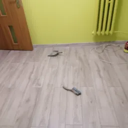 Świeżo ułożone panele podłogowe w pokoju z jasnozielonymi ścianami, widoczne narzędzia do sprzątania - zmiotka i szufelka, oraz grzejnik żeberkowy.