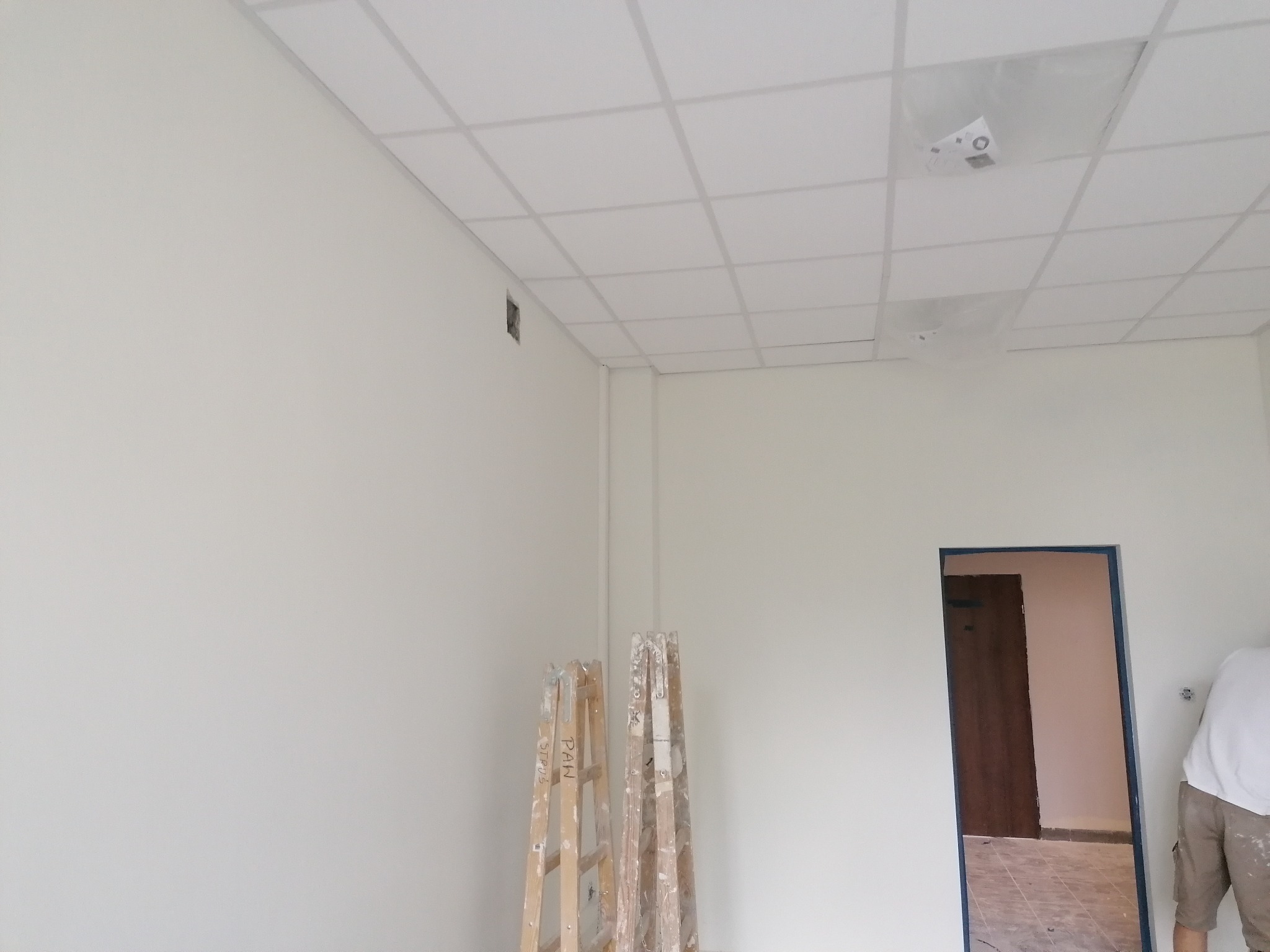 Wnętrze w trakcie malowania: białe ściany, sufit kasetonowy z folią ochronną, drabiny malarskie z napisami, widok na drzwi w sąsiednim pomieszczeniu, fragment sylwetki malarza.