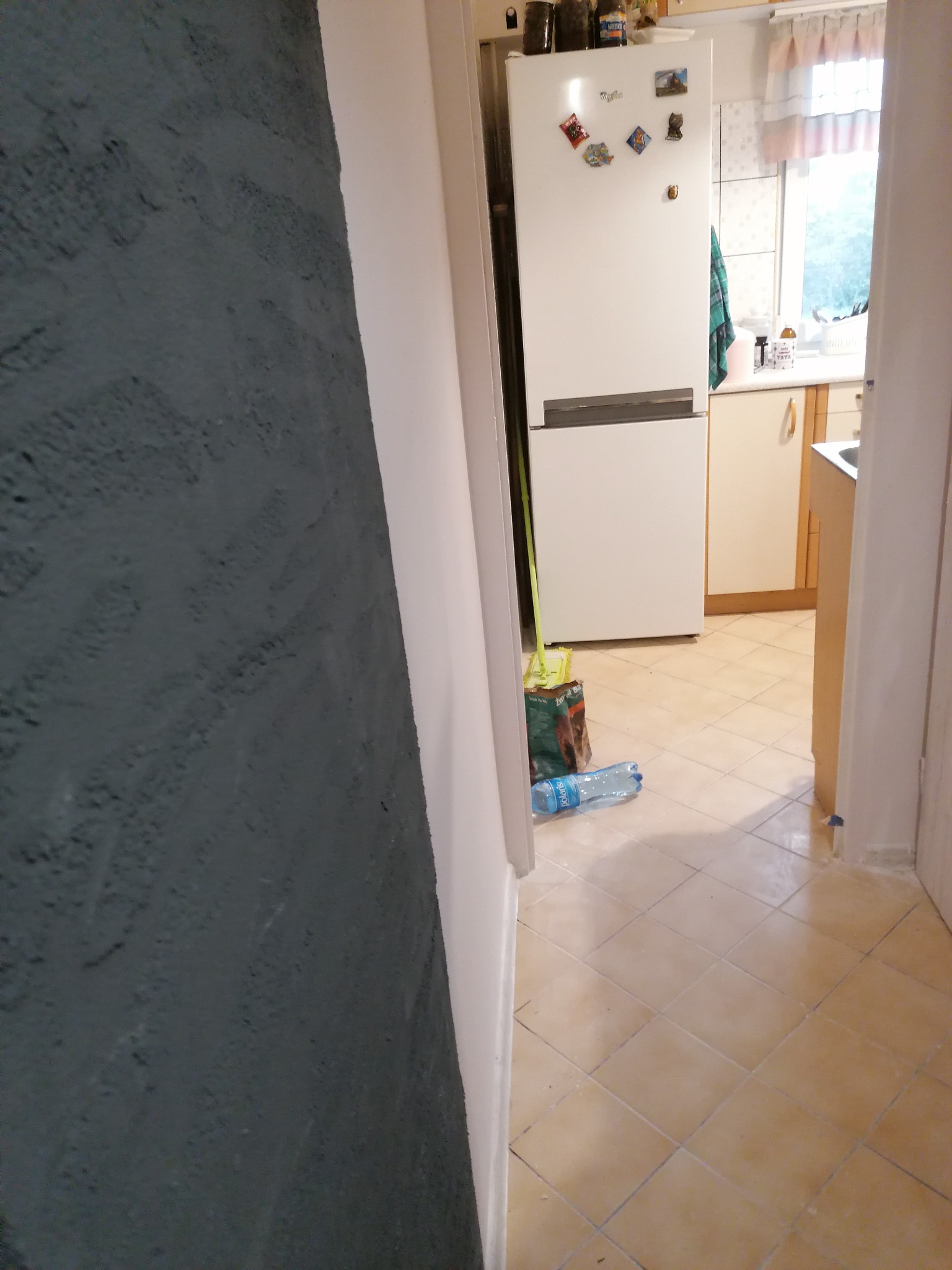 Fragment ściany z szarą, nierównomierną fakturą w tle widok na kuchnię z białą lodówką, mopem i butelką wody na podłodze z płytek w odcieniach beżu.