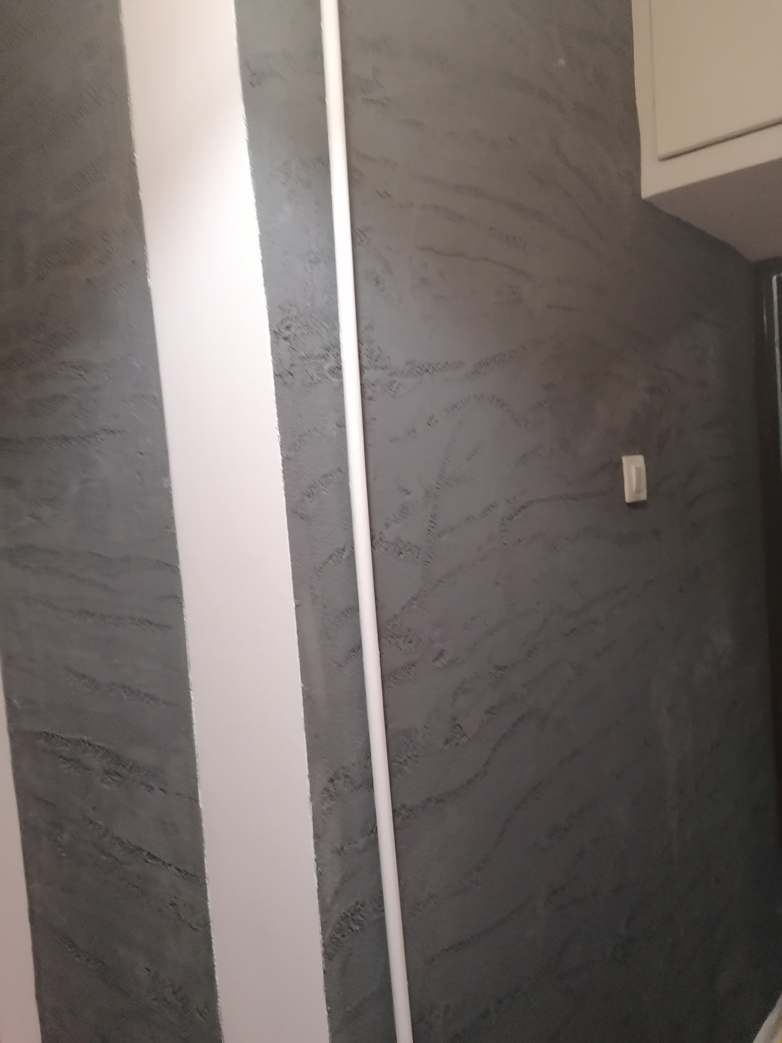 Szara ściana z dekoracyjną fakturą i białymi, pionowymi elementami wykończeniowymi, widoczny włącznik światła i fragment szafki.