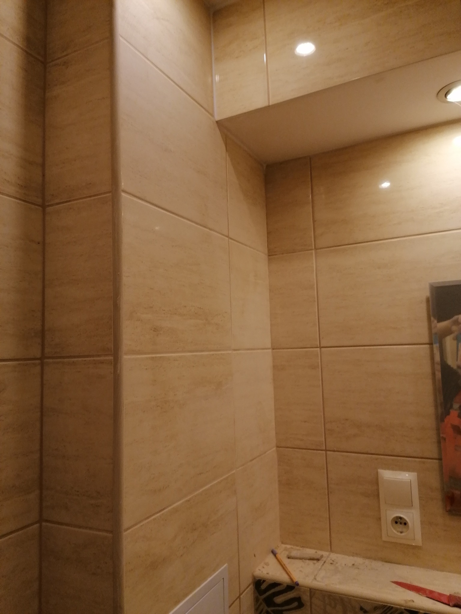 Wykończona ściana łazienki z beżowych płytek ceramicznych, widoczny włącznik światła i gniazdko elektryczne, fragment blatu z narzędziami.