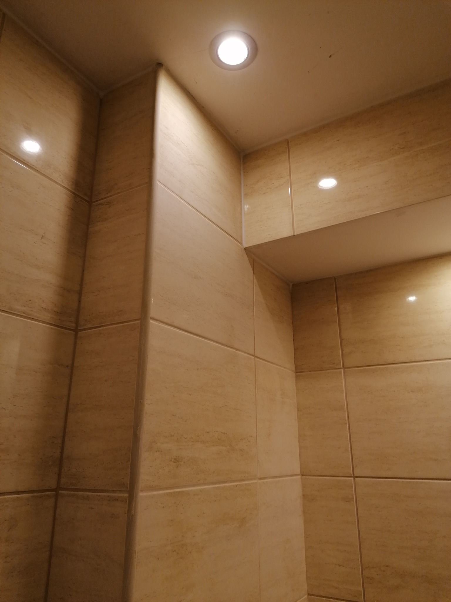 Wykończony narożnik łazienki z beżowymi płytkami imitującymi trawertyn, oświetlony trzema punktowymi lampami sufitowymi.