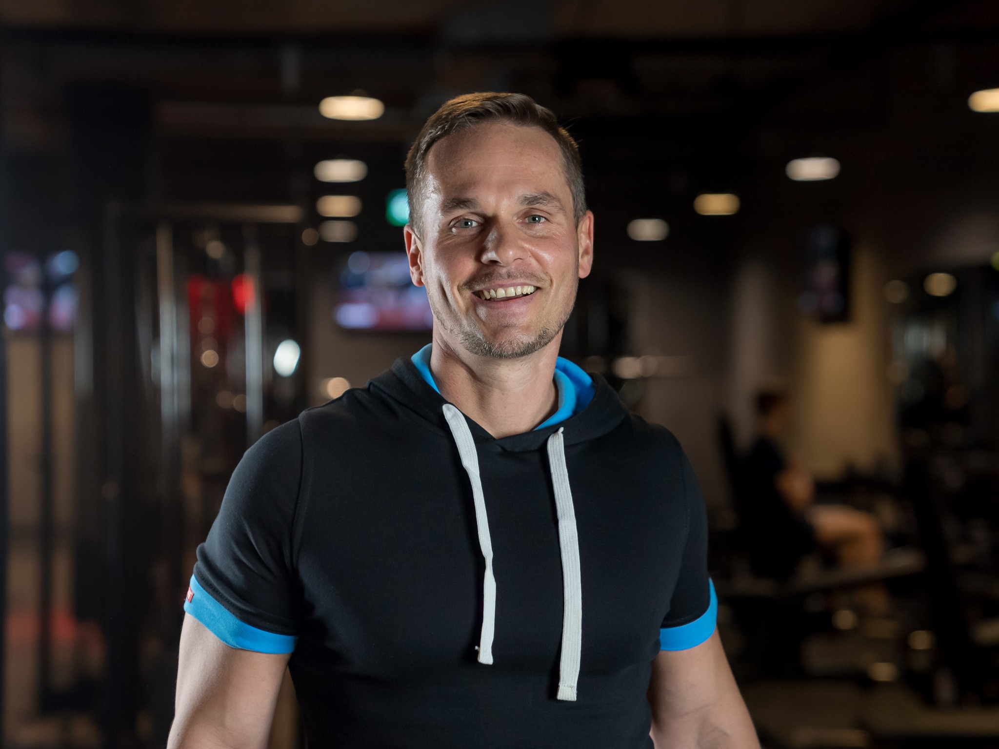 Uśmiechnięty mężczyzna w czarnej bluzie z kapturem i niebieskimi wstawkami na rękawach, stojący na tle sali gimnastycznej z widocznymi urządzeniami do ćwiczeń.