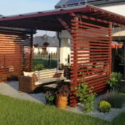 Nowoczesna, drewniana pergola z poziomymi listwami, wyposażona w rattanowy zestaw wypoczynkowy z beżowymi poduszkami i wiszące doniczki z kwiatami, przylegająca do białego domu z szarą kostką brukową.