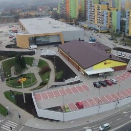 INWESTPROJEKT - Widok z góry na ukończony budynek supermarketu z parkingiem, otoczony zielenią i drogami, z blokami mieszkalnymi w tle w pochmurny dzień.