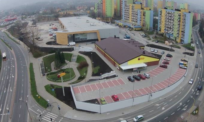 Widok z góry na ukończony budynek supermarketu z parkingiem, otoczony zielenią i drogami, z blokami mieszkalnymi w tle w pochmurny dzień.