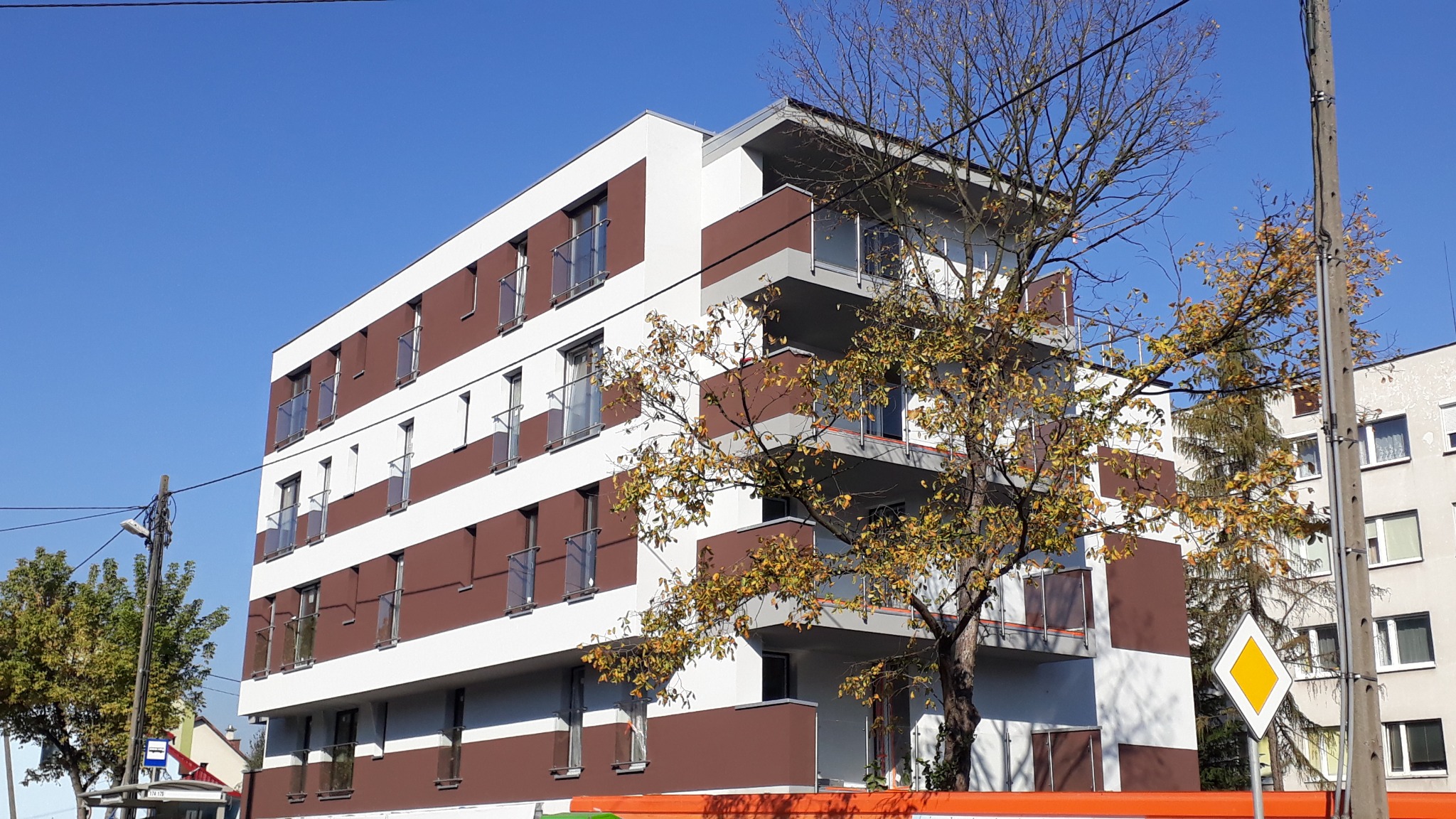 Nowoczesny budynek wielorodzinny z brązowo-białą elewacją i szklanymi balkonami, sfotografowany w słoneczny dzień z perspektywy ulicy, z widocznym drzewem z żółtymi liśćmi na pierwszym planie.