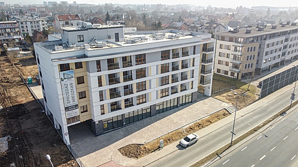 Nowoczesny, czteropiętrowy budynek mieszkalny z balkonami i dużymi oknami, z banerem reklamowym 'Sprzedaż Mieszkań' na elewacji, widziany z góry, z samochodem osobowym parkującym na ulicy.