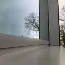 Szary profil okienny z widocznym odbiciem drzewa w szybie, zewnętrzny widok na drzewa i fragment zieleni.