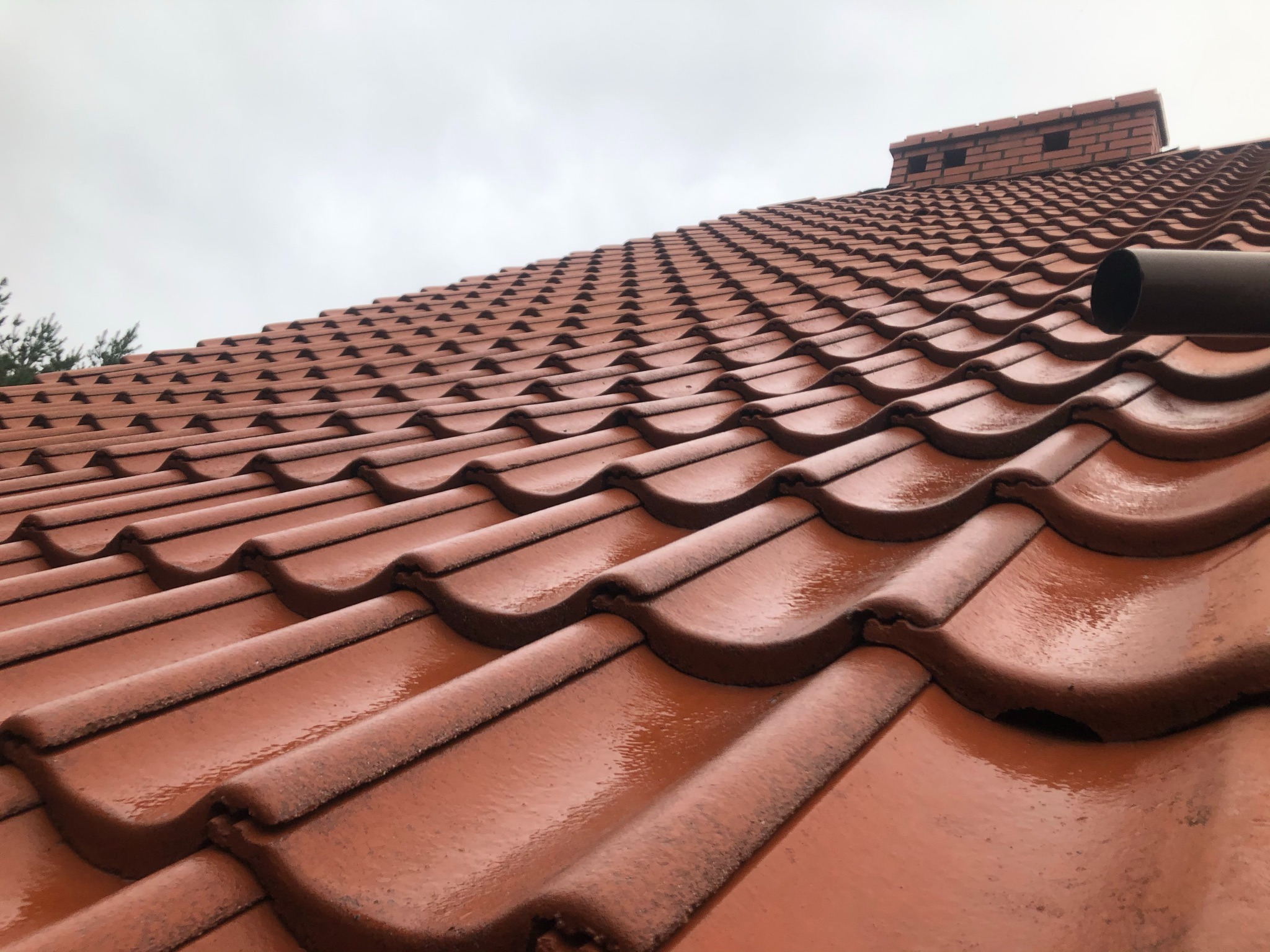 Czerwona dachówka ceramiczna, mokra po deszczu, z widocznym kominem z cegły w tle i fragmentem rynny.