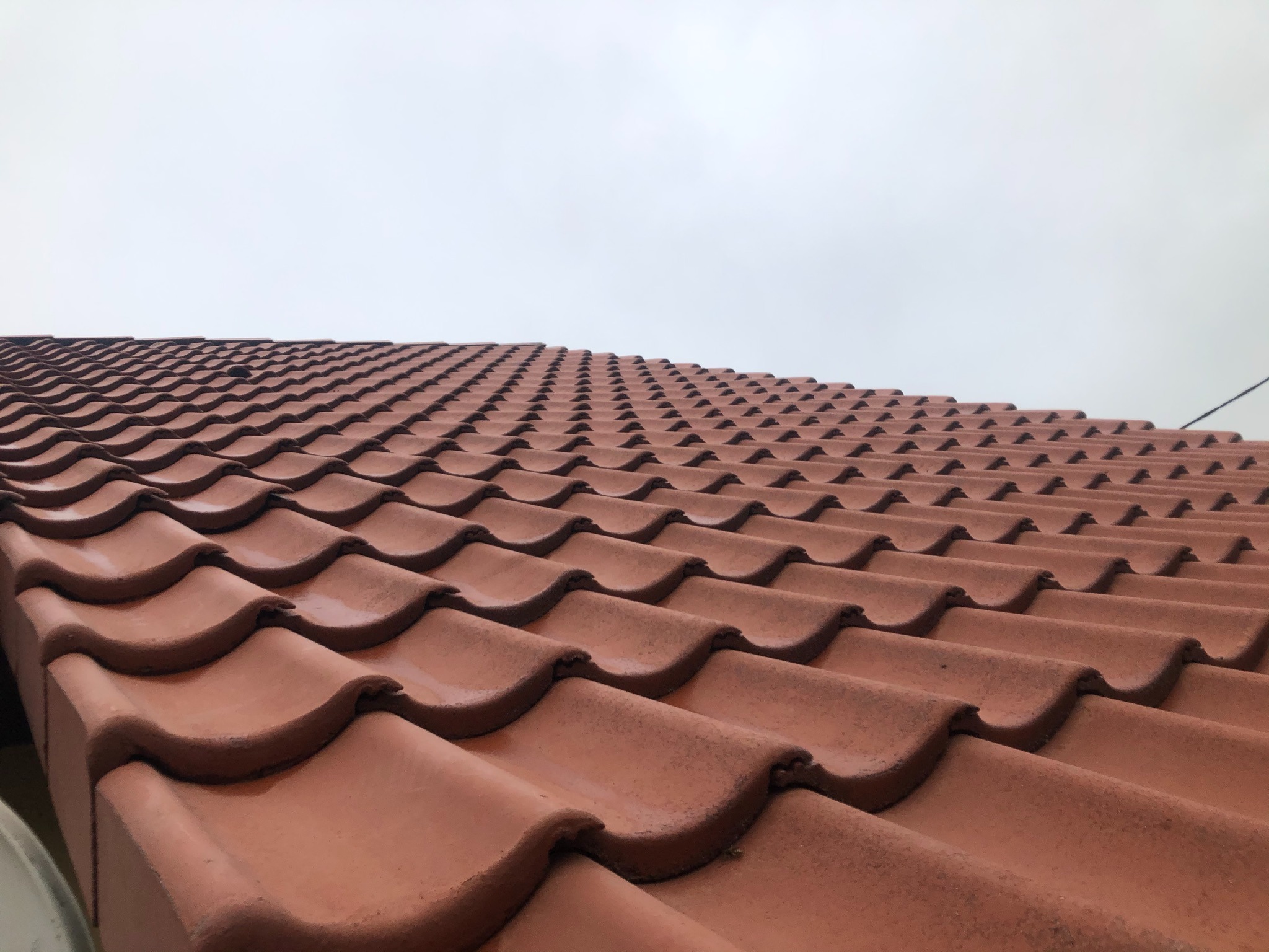 Równomiernie ułożone, lekko wilgotne dachówki ceramiczne w odcieniach terakoty, sfotografowane pod kątem, z szarym, pochmurnym niebem w tle.