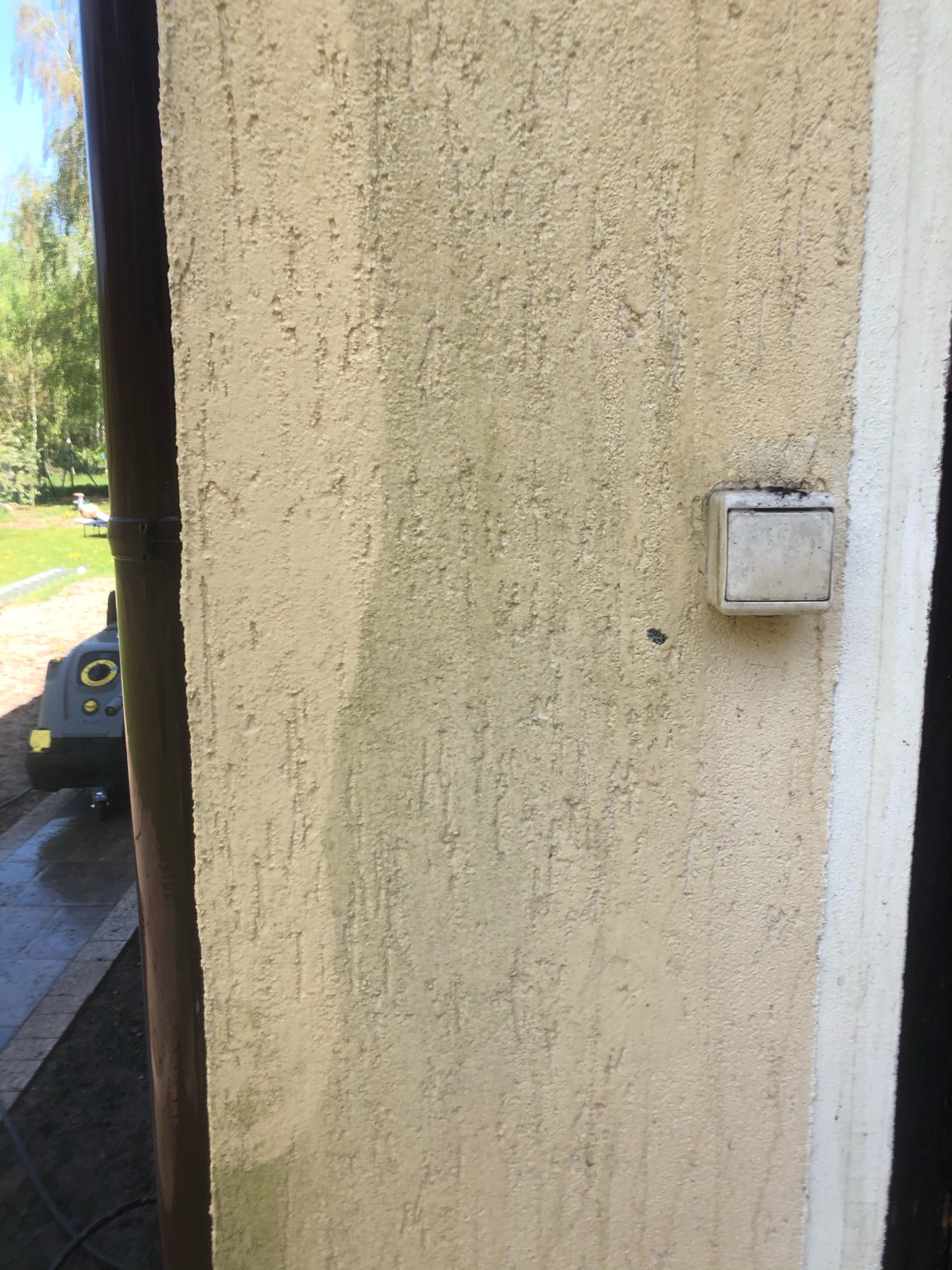 Fragment elewacji z widoczną różnicą w czystości po umyciu, z brązową rynną po lewej stronie i włącznikiem światła na ścianie, w tle trawnik i drzewa.
