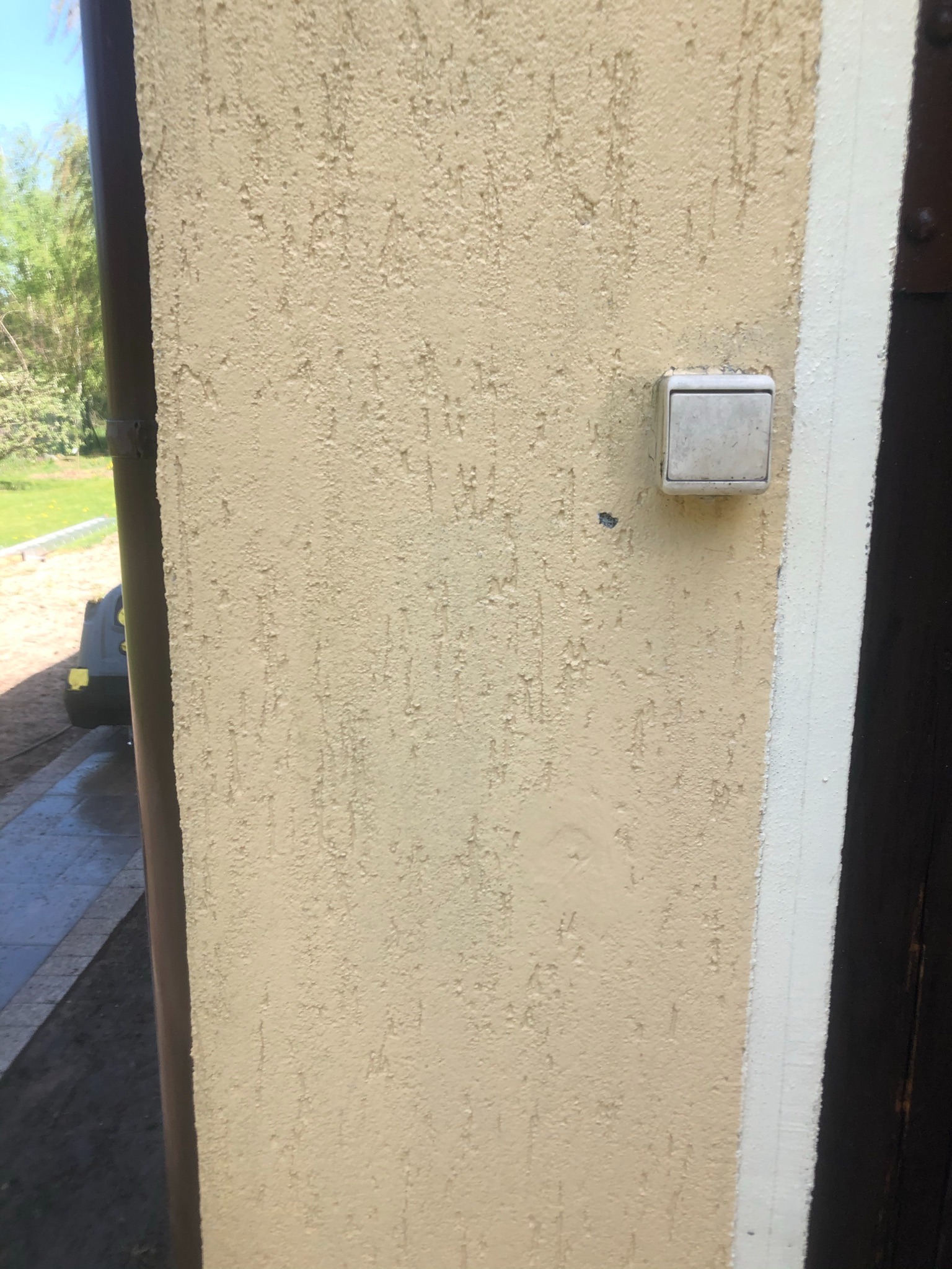 Jasnobeżowa ściana z fakturą baranka, obok brązowe drzwi z białą listwą, widoczny fragment rynny i włącznik światła. W tle zieleń trawnika i drzew.