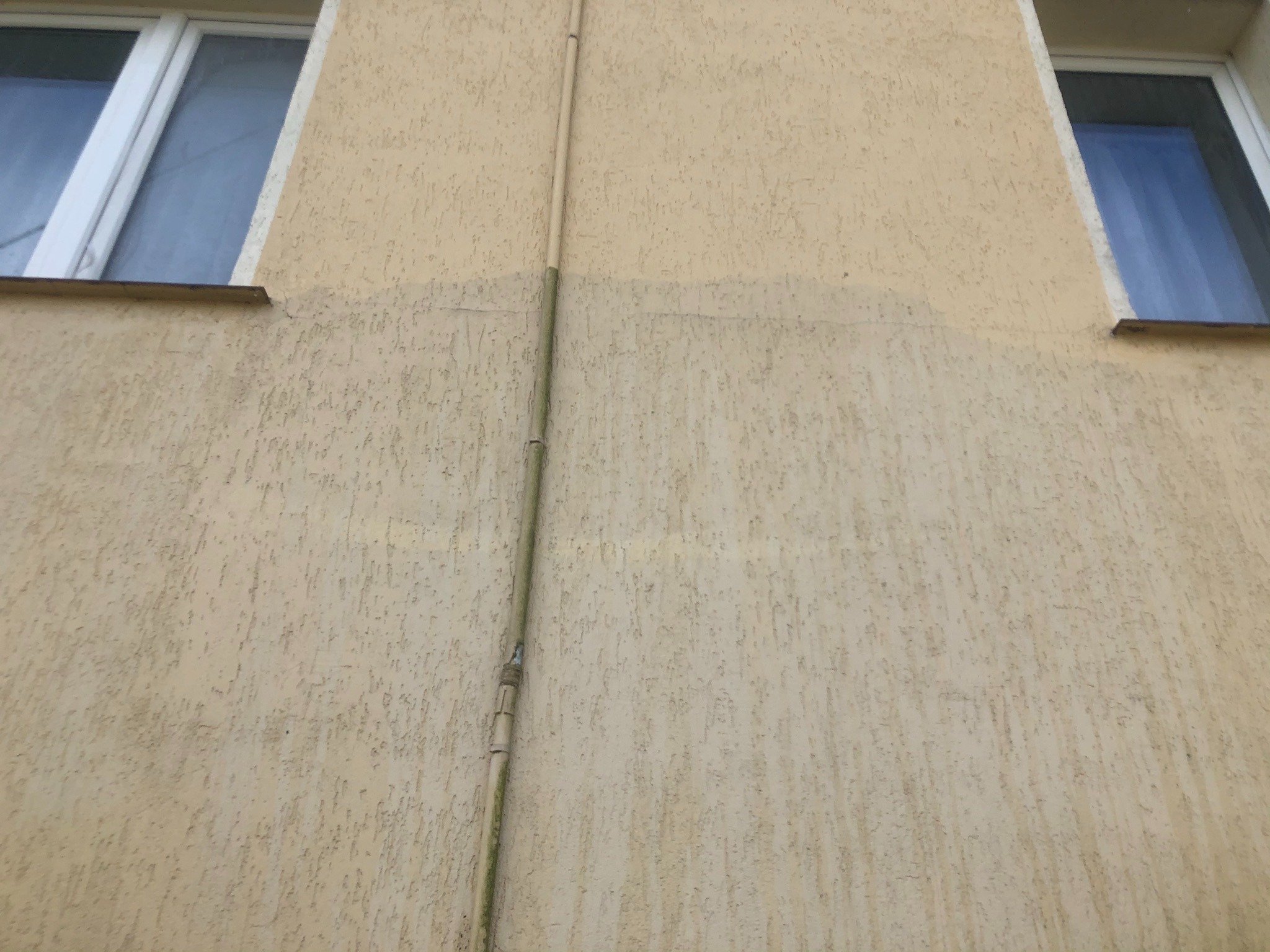 Fragment elewacji budynku z widoczną różnicą w czystości, gdzie górna część jest jaśniejsza, a dolna ciemniejsza. Pionowa rura biegnie wzdłuż ściany, częściowo pokryta mchem. Dwa okna z białymi...