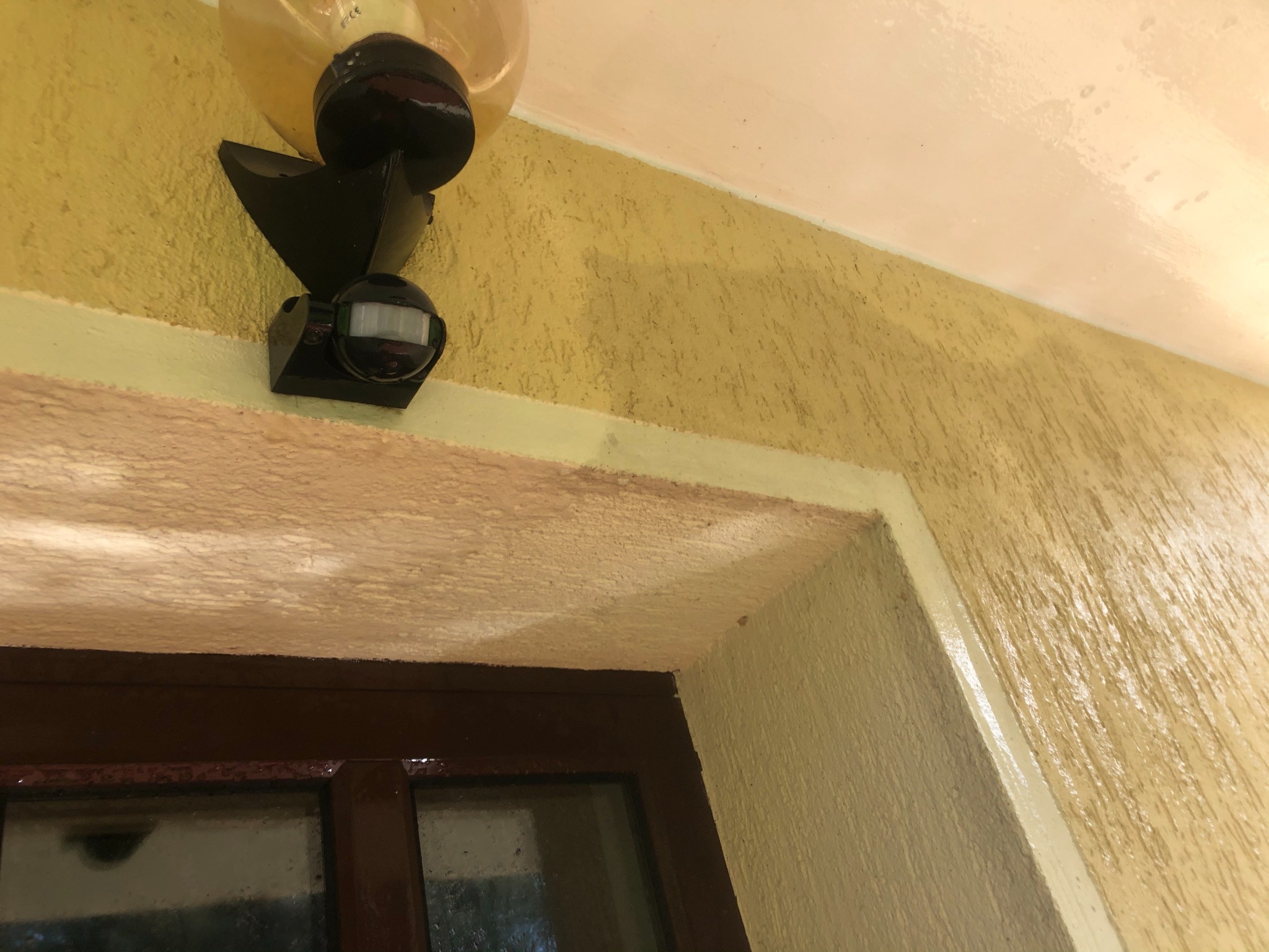 Żółta elewacja z czujnikiem ruchu i lampą nad brązową ramą okienną, widoczne niedociągnięcia malarskie.
