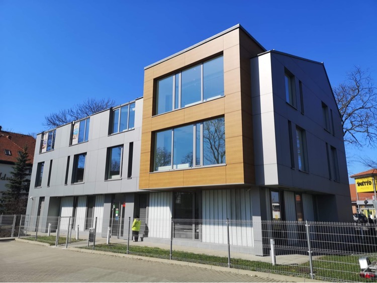 Nowoczesny budynek biurowy z elewacją w odcieniach szarości i drewna, widoczny znak Netto w tle, osoba w żółtej kurtce przed wejściem.