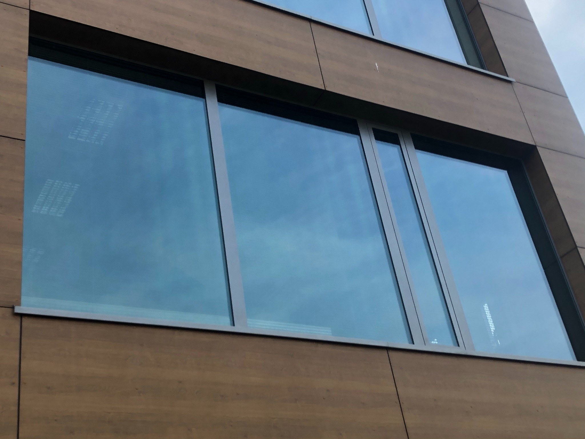 Nowoczesna elewacja budynku z drewnianymi panelami i dużymi, czystymi oknami, w których odbija się błękitne niebo i fragmenty sąsiednich budynków.
