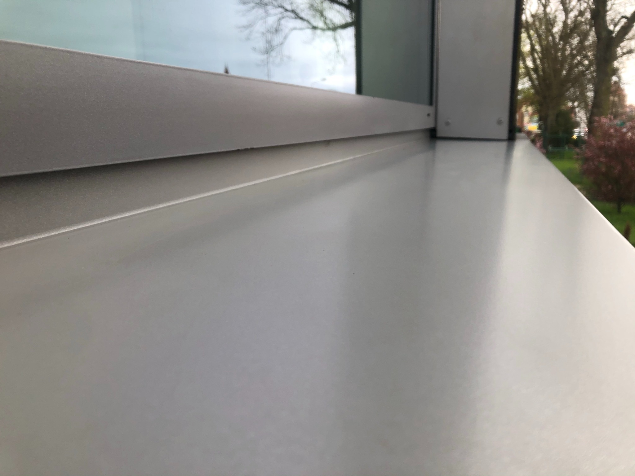 Szary parapet zewnętrzny z widocznym oknem i fragmentem zieleni w tle, uchwycony z niskiej perspektywy.