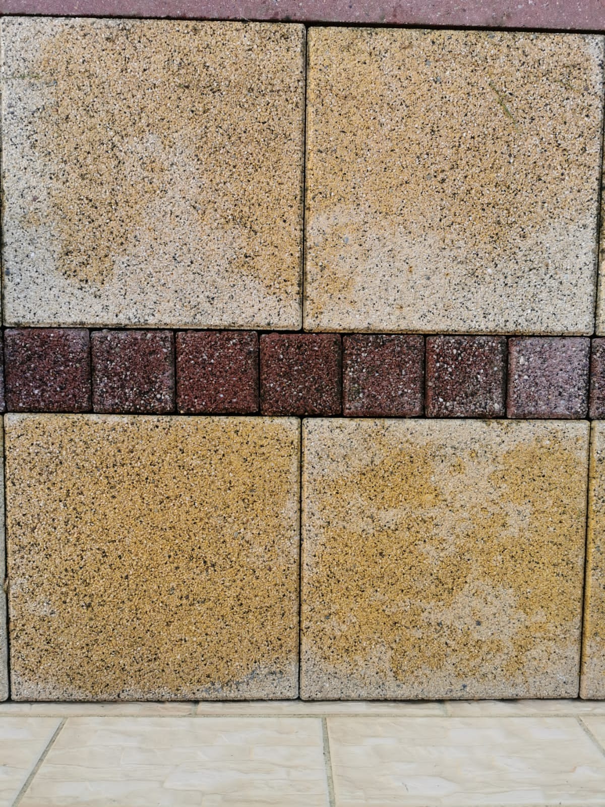 Fragment elewacji budynku z żółtych bloczków z ciemnoczerwonym pasem bloczków dekoracyjnych, widoczny dolny fragment ściany i podłoga z beżowych płytek.