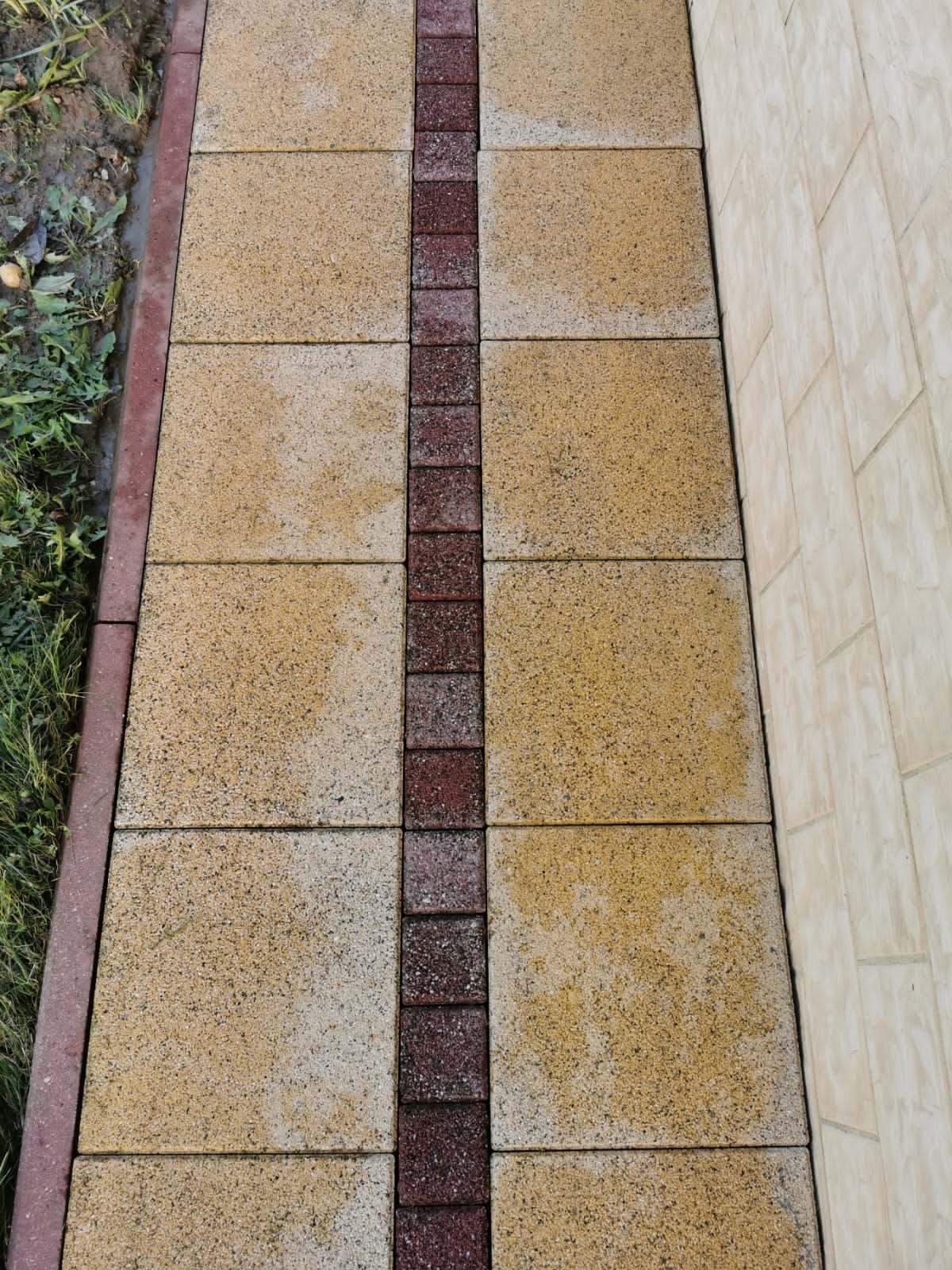 Ułożona ścieżka z beżowych płyt chodnikowych z bordowym pasem pośrodku, widok z góry, obok ściana budynku i trawa.