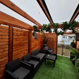 Zadaszony taras z drewnianą konstrukcją, sztuczną trawą i meblami ogrodowymi. Dekoracje roślinne wiszą na belkach, tworząc przytulną przestrzeń.