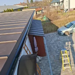 Widok z góry na brązowy dach z blachy trapezowej, z widoczną rynną, drabiny i fragment posesji z kostką brukową oraz budynkiem gospodarczym w słoneczny dzień.