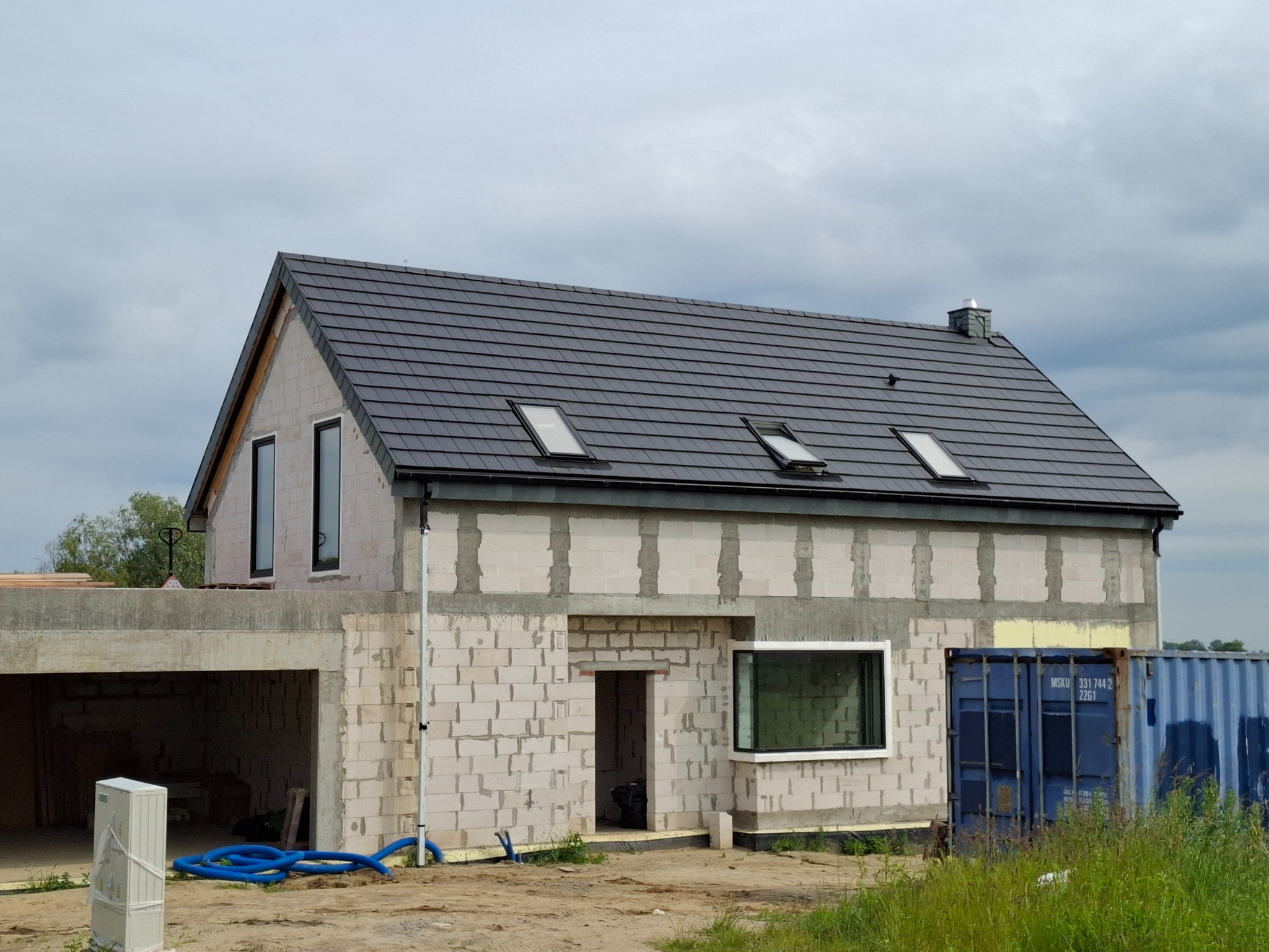 Dom w stanie surowym zamkniętym z ciemną dachówką i oknami. Widoczne bloczki betonowe, garaż w bryle budynku i kontener. Ujęcie z poziomu gruntu.