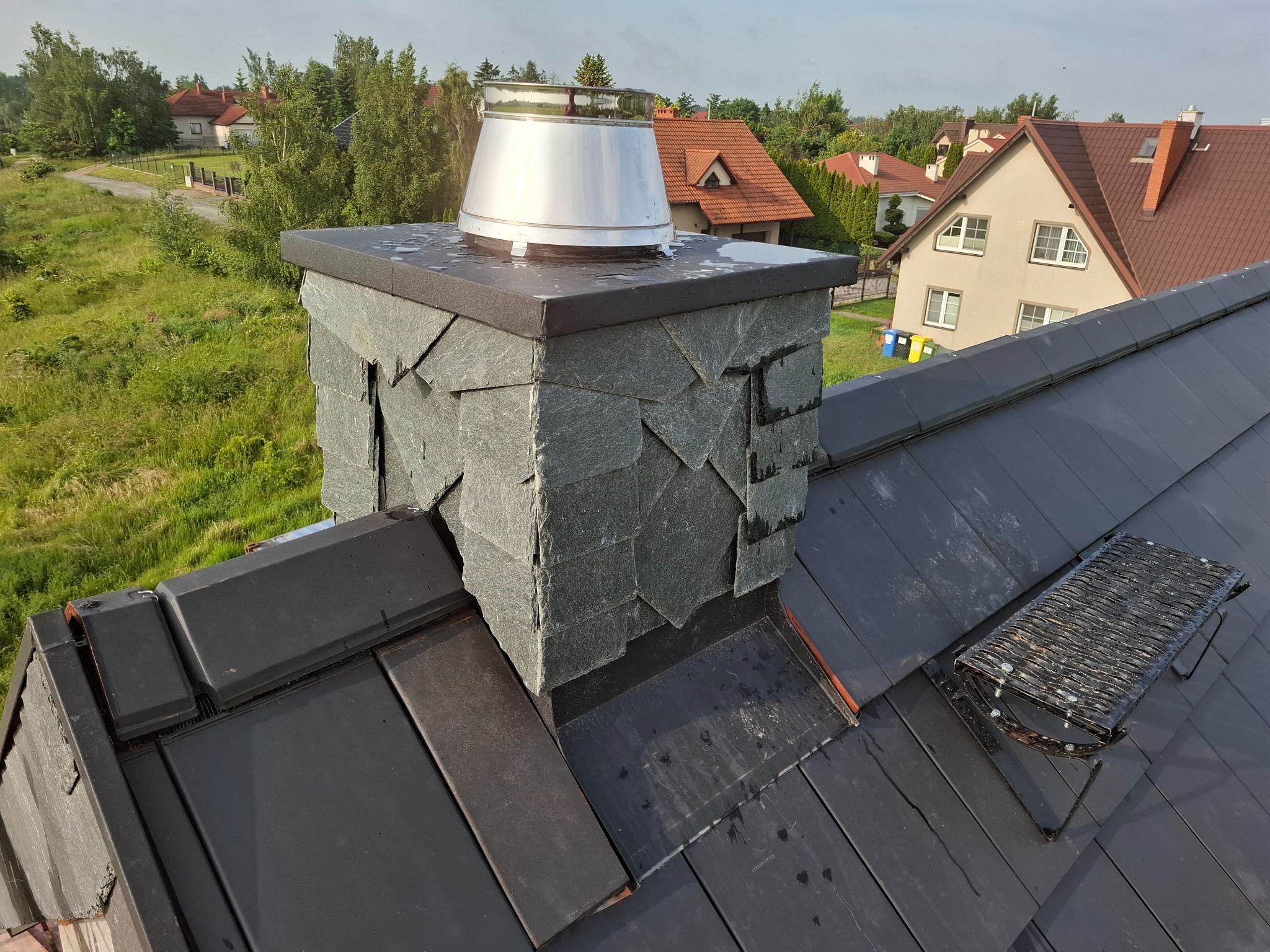 Wykończony komin z szarego kamienia łupkowego zwieńczony metalowym daszkiem na ciemnym dachu z dachówki ceramicznej, w tle zabudowa jednorodzinna i zieleń.