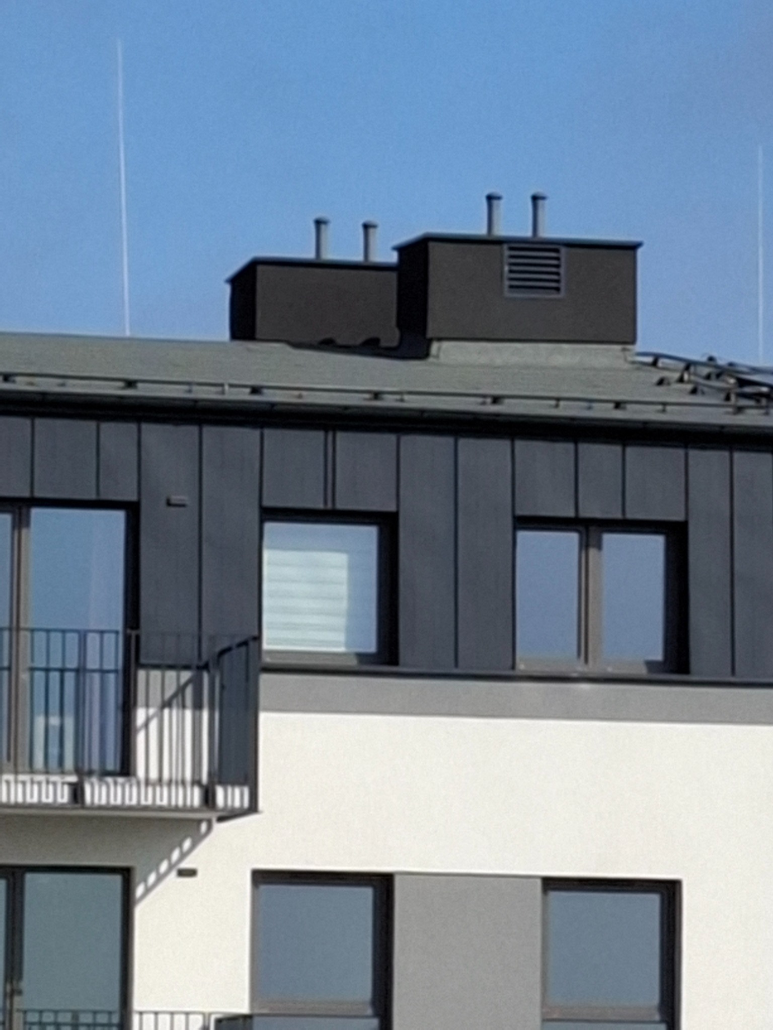 Elewacja budynku z szarego panelu z balkonem. Na dachu widoczne kominy wentylacyjne. Minimalistyczna, nowoczesna architektura.