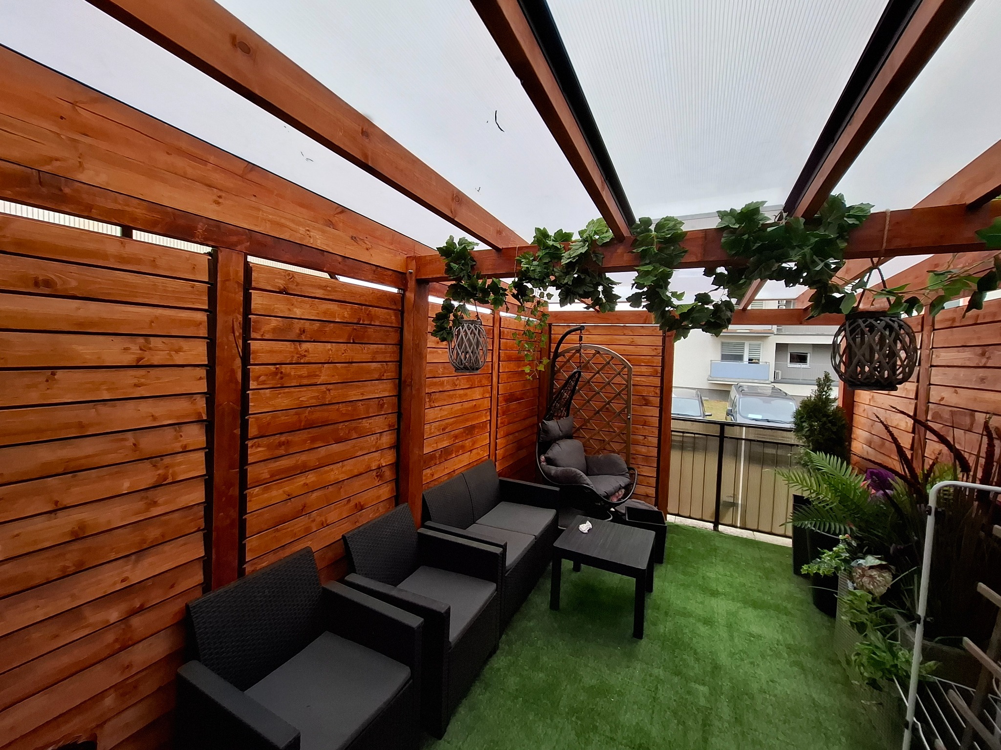 Zadaszony taras z drewnianą konstrukcją, sztuczną trawą i meblami ogrodowymi. Dekoracje roślinne wiszą na belkach, tworząc przytulną przestrzeń.