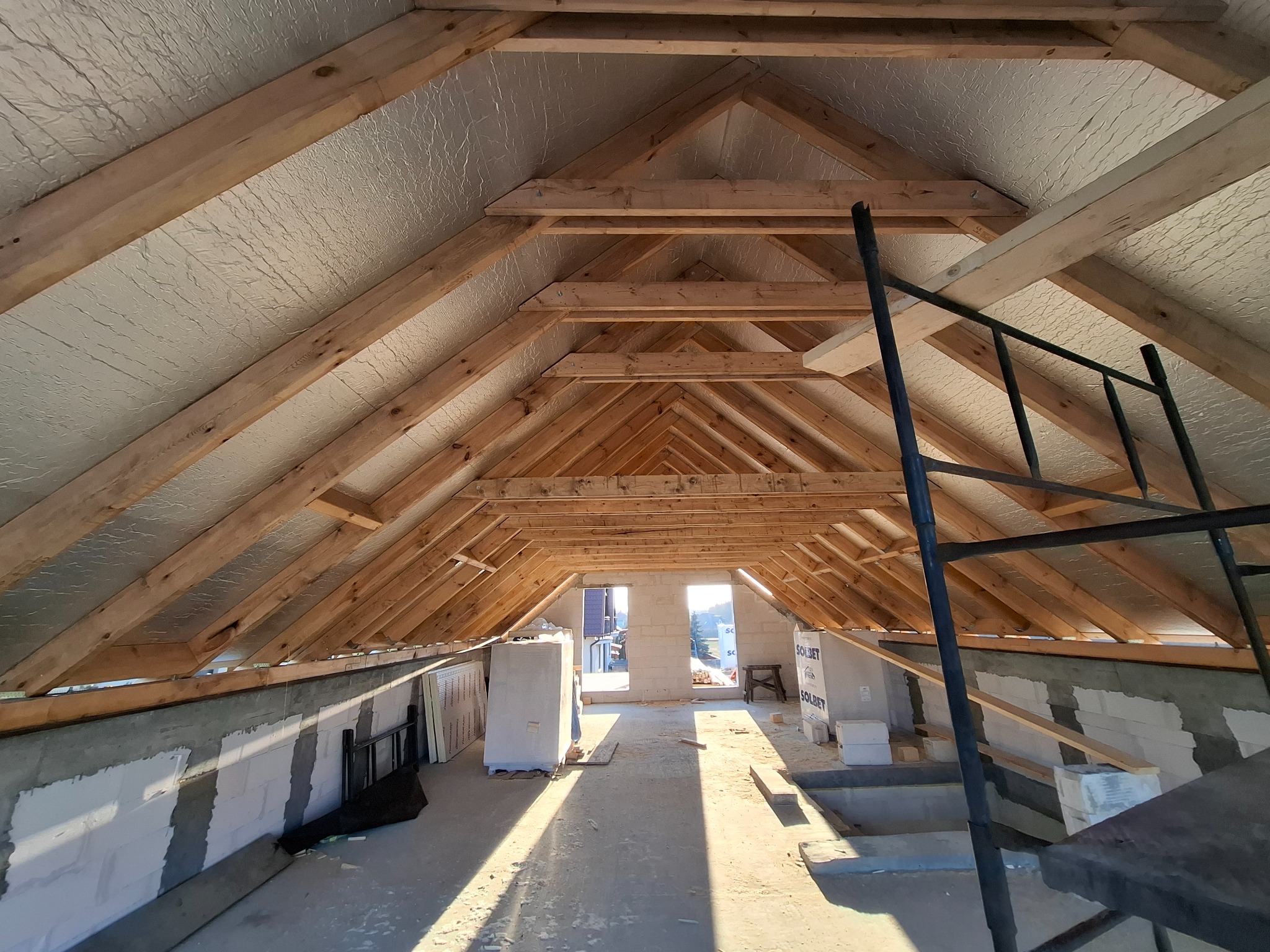 Ażurowa konstrukcja drewnianej więźby dachowej z ociepleniem w budynku w trakcie budowy. W tle okna w murze i fragment drabiny.