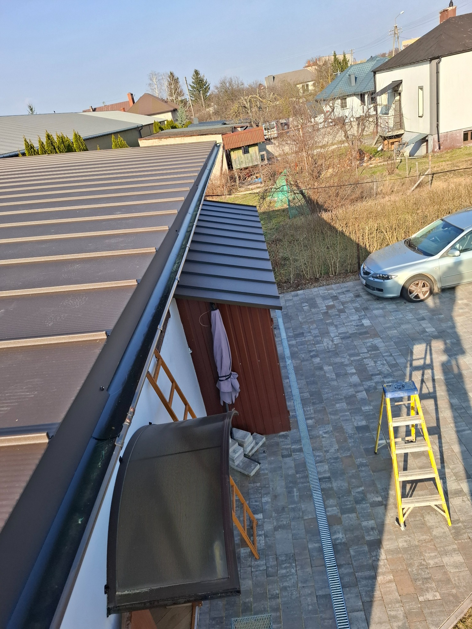 Widok z góry na brązowy dach z blachy trapezowej, z widoczną rynną, drabiny i fragment posesji z kostką brukową oraz budynkiem gospodarczym w słoneczny dzień.