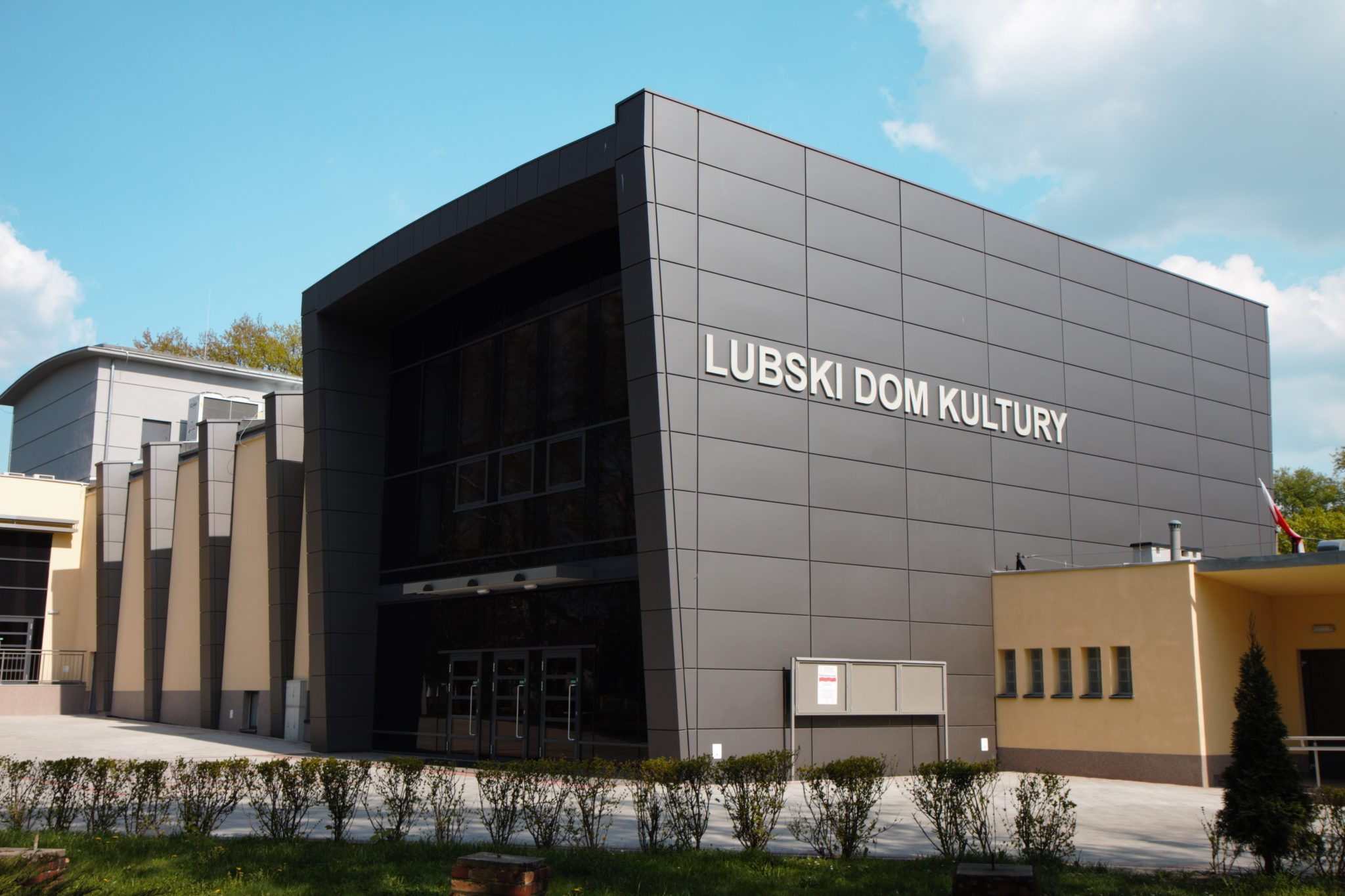 Budynek Lubskiego Domu Kultury z nowoczesną elewacją w odcieniach szarości, z dużymi przeszkleniami i minimalistycznym napisem, otoczony niskimi krzewami i trawnikiem.