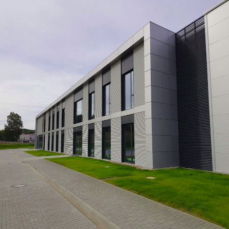 Nowoczesny budynek biurowy z elewacją z paneli aluminiowych w odcieniach szarości, widok z poziomu chodnika, z zielonym trawnikiem przy fasadzie.